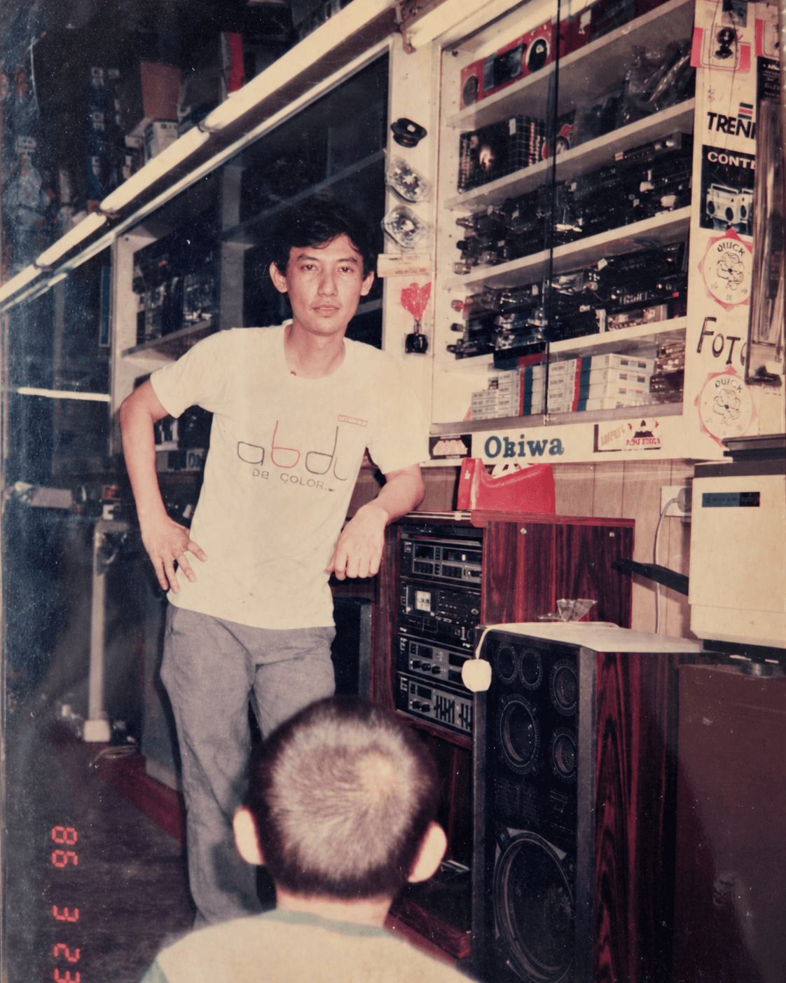 陈瑞发 1986 年在哥打丁宜店里的音响柜台前 — 穿着米色 'abdj' T 恤、靠在一叠音响器材上，前景可以看到一个孩子的后脑勺。