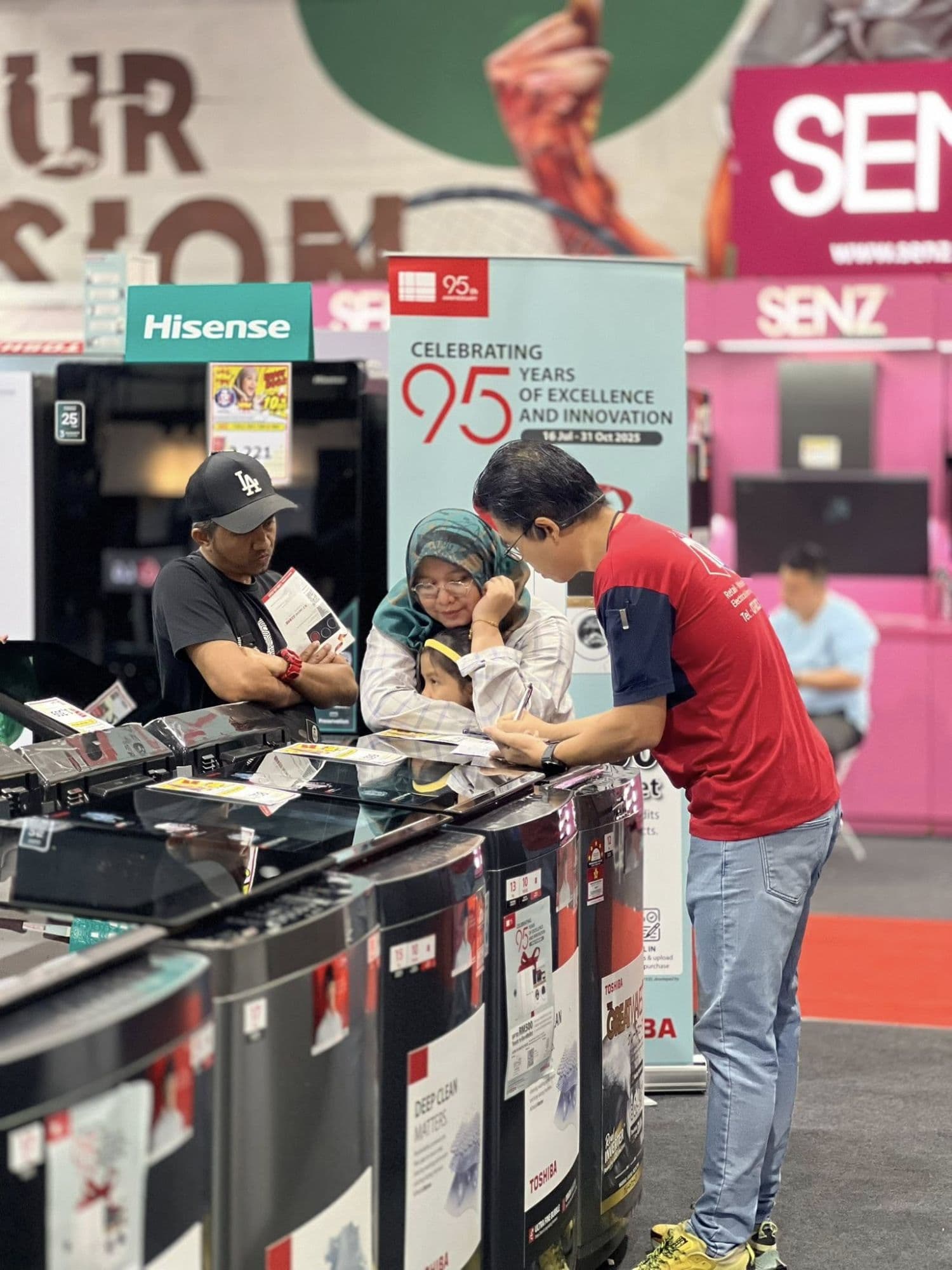 A Tanradio sales associate writes up a quote for a Malay family — father, mother in hijab, and child — at the Toshiba 95th anniversary event.